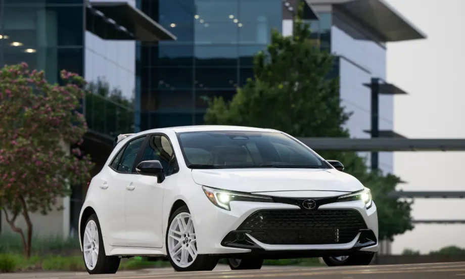 An Ice Cap Toyota Corolla Hatchback drives on the road with a glass building behind it.