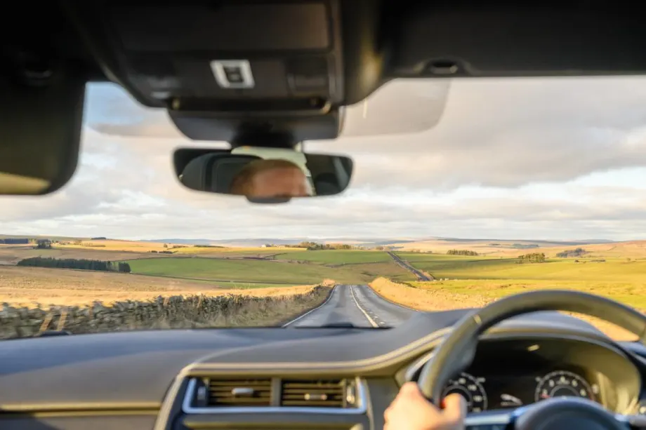 A car renter driving a right-side steering wheel on the left side of a two-lane road.