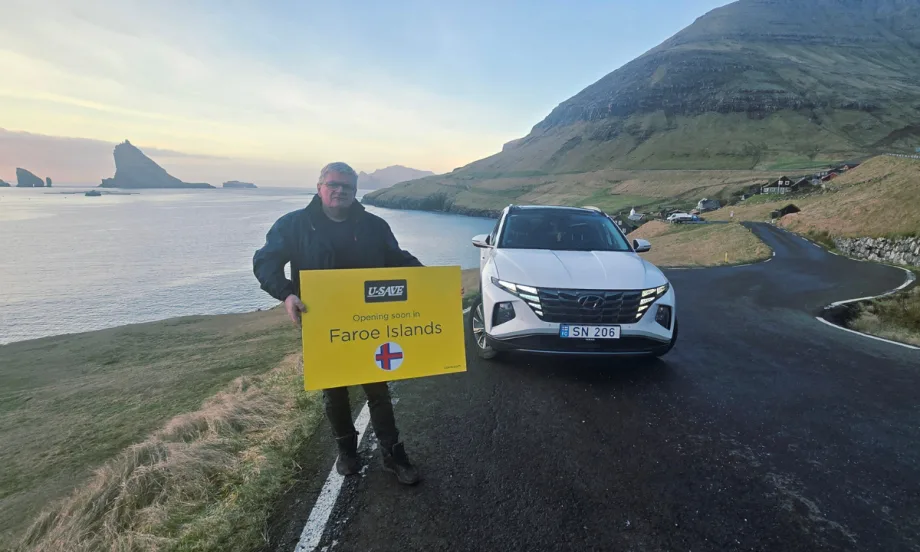 U-Save franchisee holding a promotional sign in front of a white rental car along the rugged coastline of the Faroe Islands.