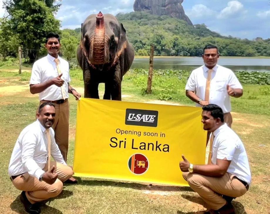 Rental car franchise employees hold up a yellow square U-Save banner in front of a rural scene featuring a protruding mountain, a distant lake, and a green field.