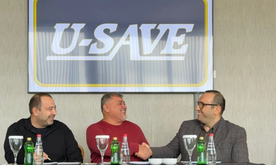 Three U-Save franchise operators are congratulating each other along a horizontal table beneath a large framed U-Save logo print.