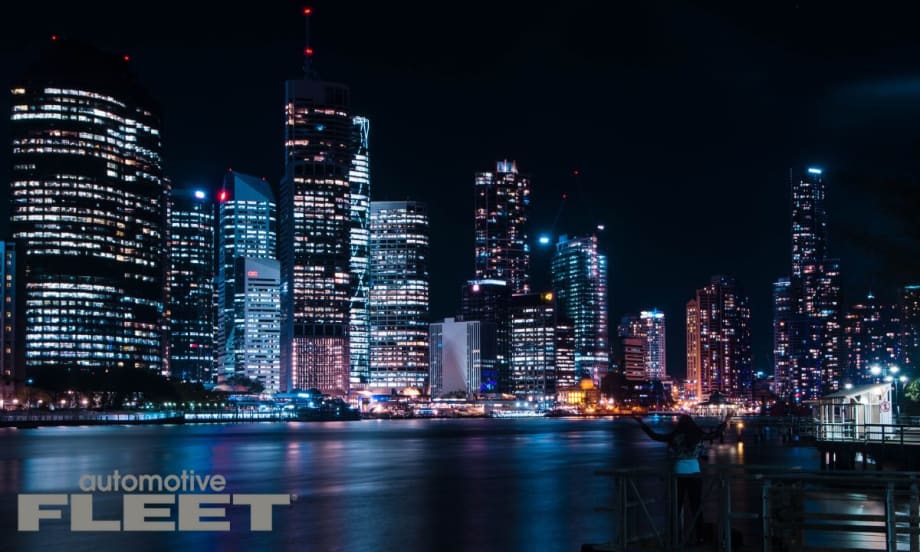 Nighttime view of a modern city skyline reflected in the water, with brightly lit skyscrapers and a silhouetted figure standing on a dock in the foreground.
